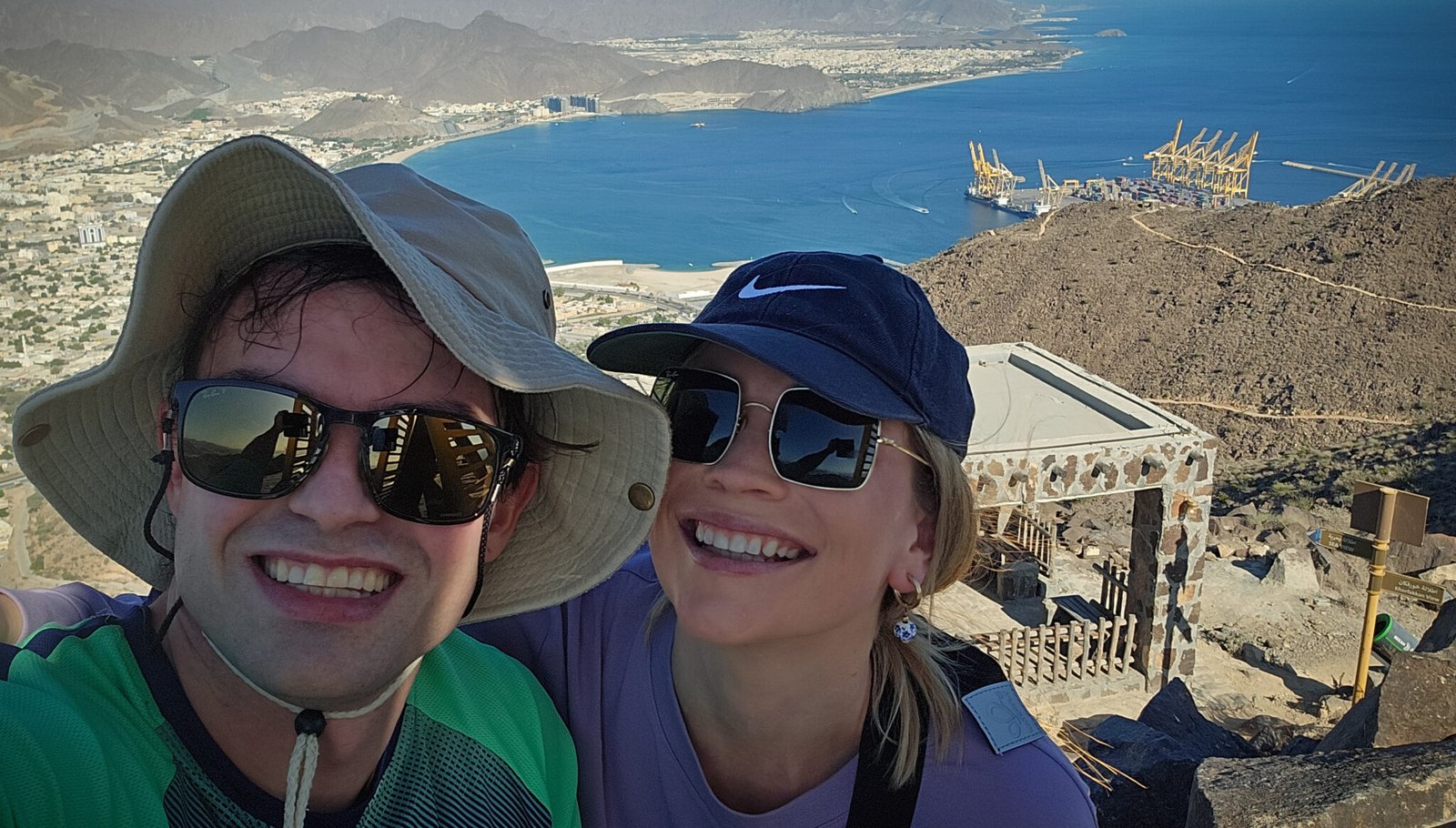 Hikers enjoying panoramic views of Khorfakkan Bay, port facilities, and Hajar Mountains from the Al Rabi Mountain summit in UAE.