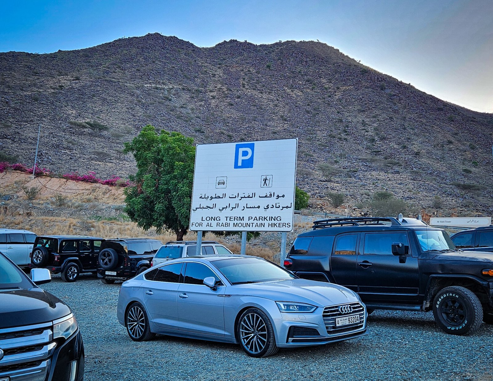 Long-term parking lot for Al Rabi Mountain hikers filled with vehicles beneath rocky hillside with bilingual parking sign and morning blue sky.