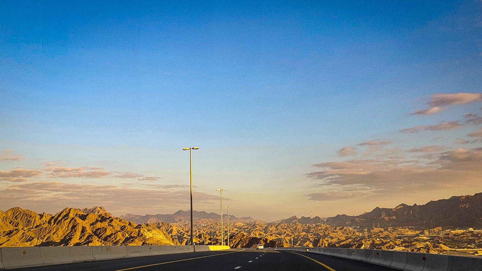 Highway S142 cutting through golden-lit Hajar Mountains during sunrise with clear blue sky and safety barriers visible.
