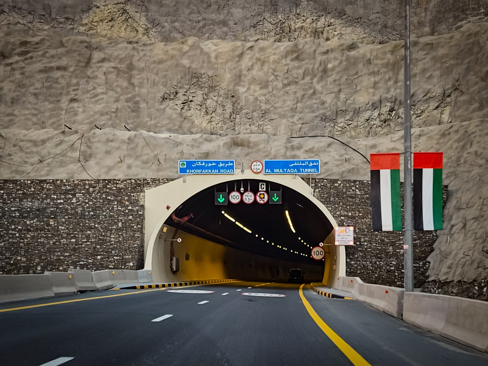 Entrance to Al Multaqa Tunnel on Khorfakkan Road with bilingual signage, safety barriers, and UAE flag displayed on nearby pole.