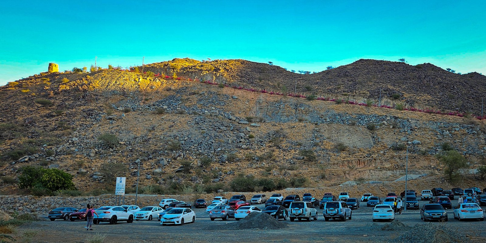Crowded parking area below Al Rabi Mountain with numerous vehicles and the historic watchtower visible on the hilltop, illuminated in the morning light under a turquoise sky.