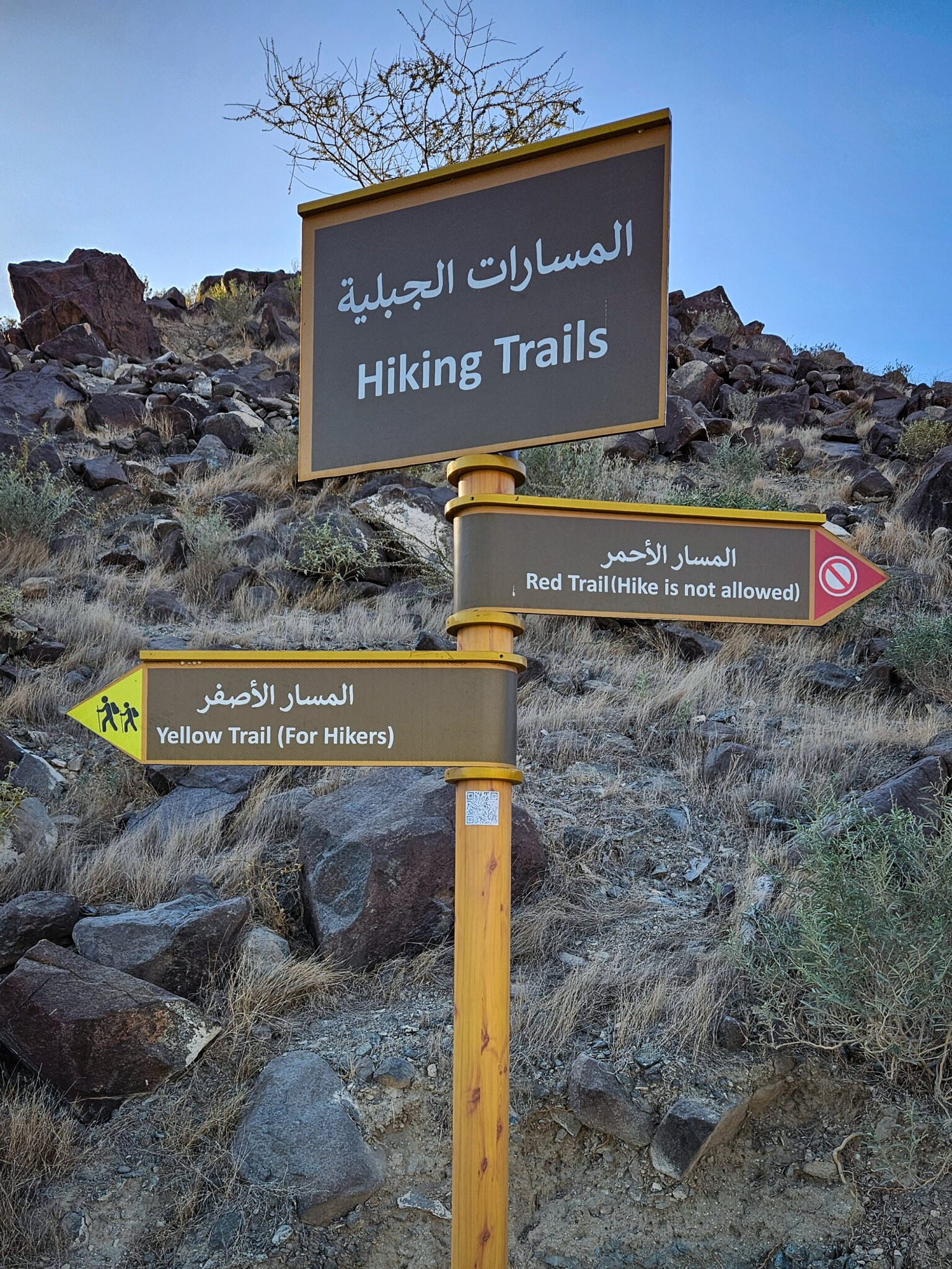 Bilingual hiking trail signpost on the Al Rabi hiking trail showing Yellow Trail for hikers and Red Trail where hiking is not allowed against a rocky mountain backdrop.
