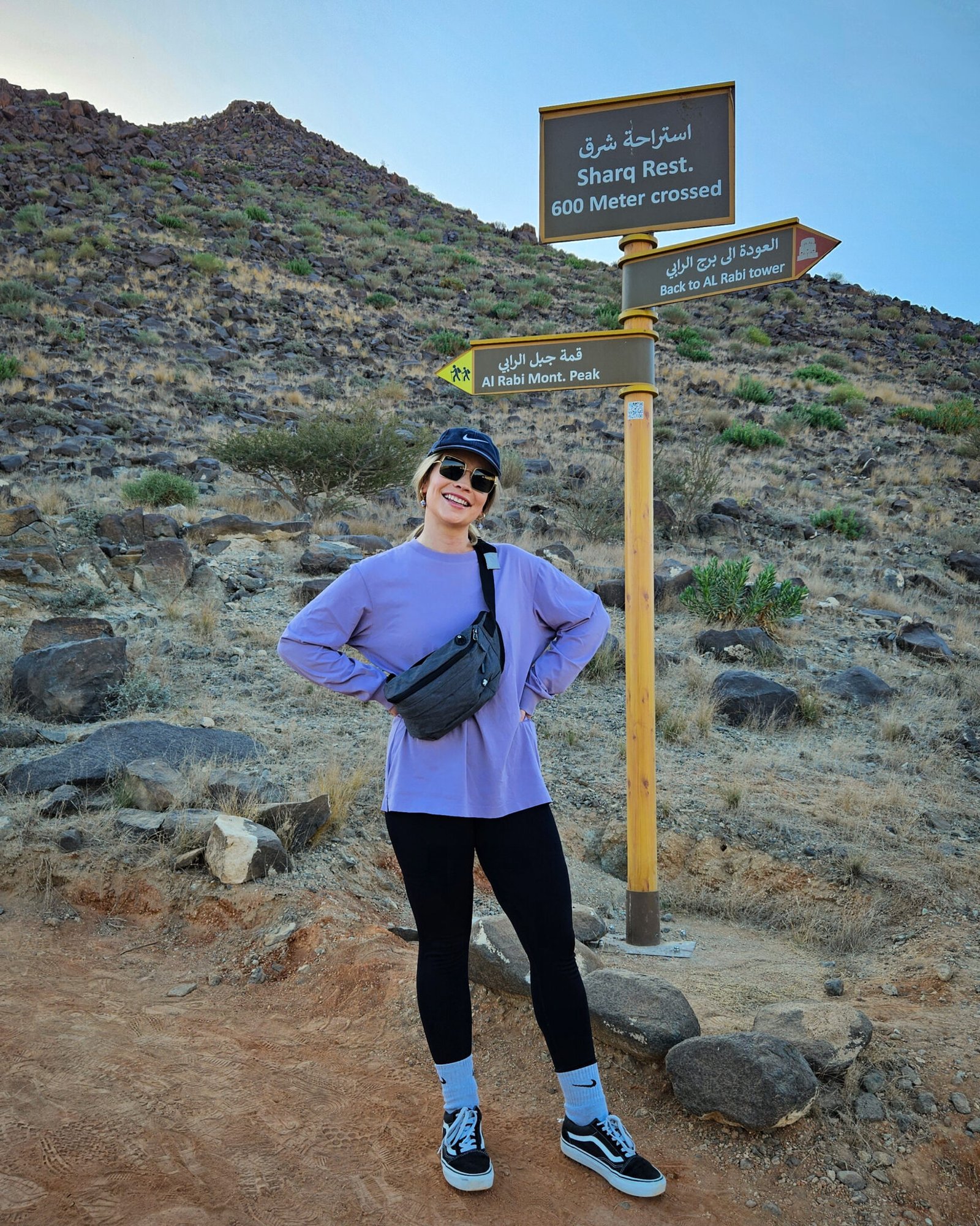 Woman in purple top and black leggings standing beside trail signpost showing directions to Al Rabi Mountain Peak and back to the tower.
