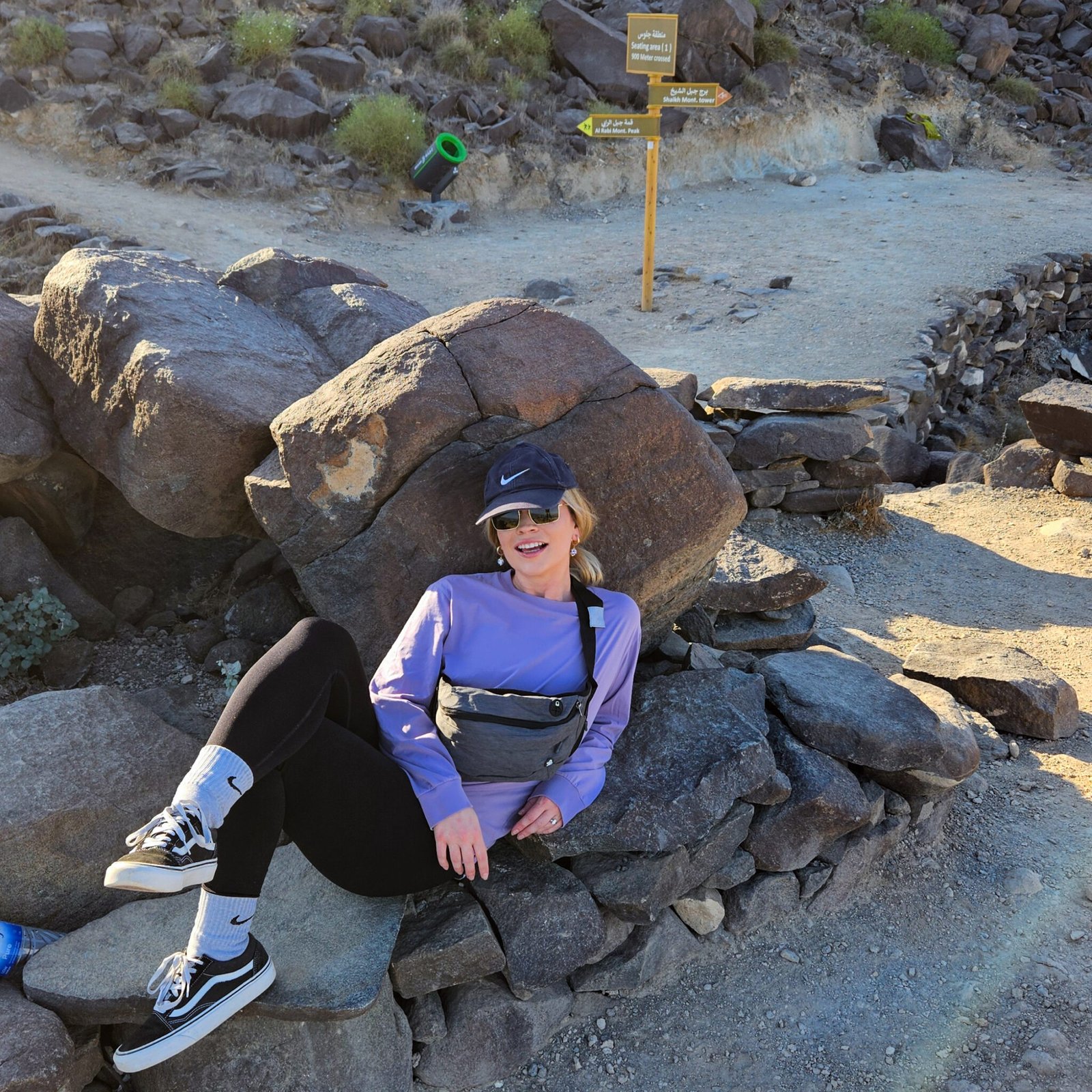Woman in purple top sitting on rocks at a trail junction with wooden directional signs visible in the background.