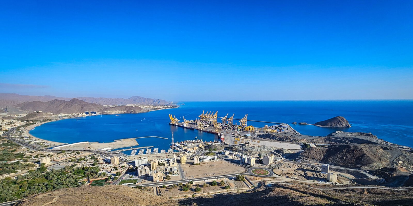 Panoramic view of Khorfakkan Bay with port facilities, blue waters, Shark Island, and Hajar Mountains from elevated trail viewpoint.
