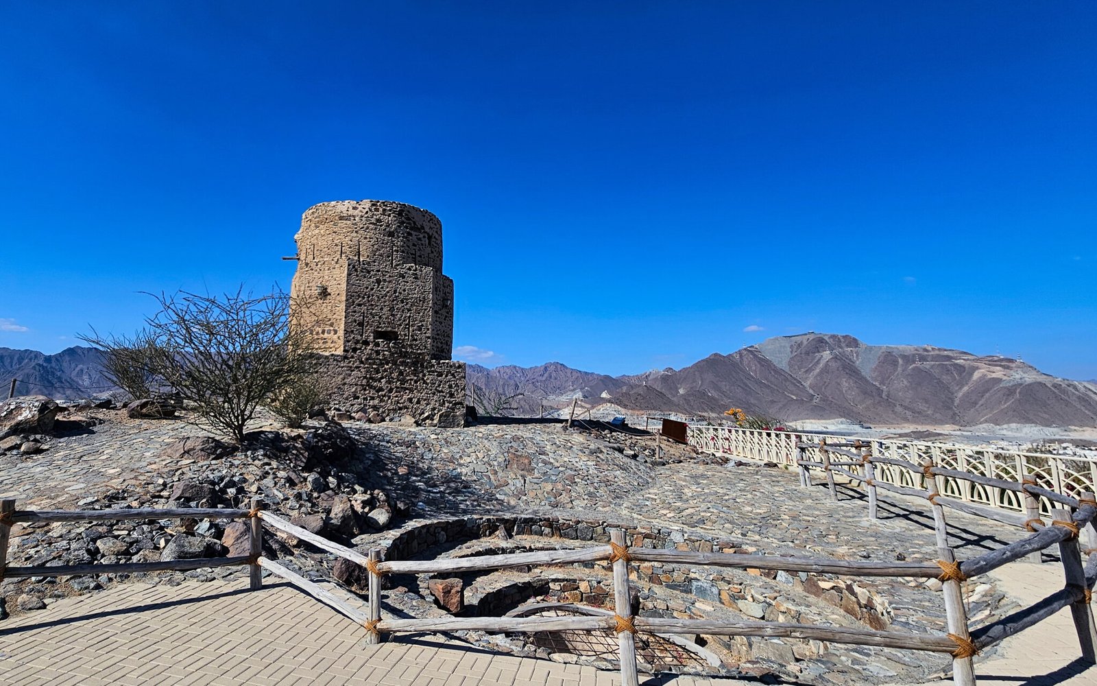 Historical Al Rabi watchtower at the hiking trail starting point surrounded by protective wooden fencing with mountain backdrop.