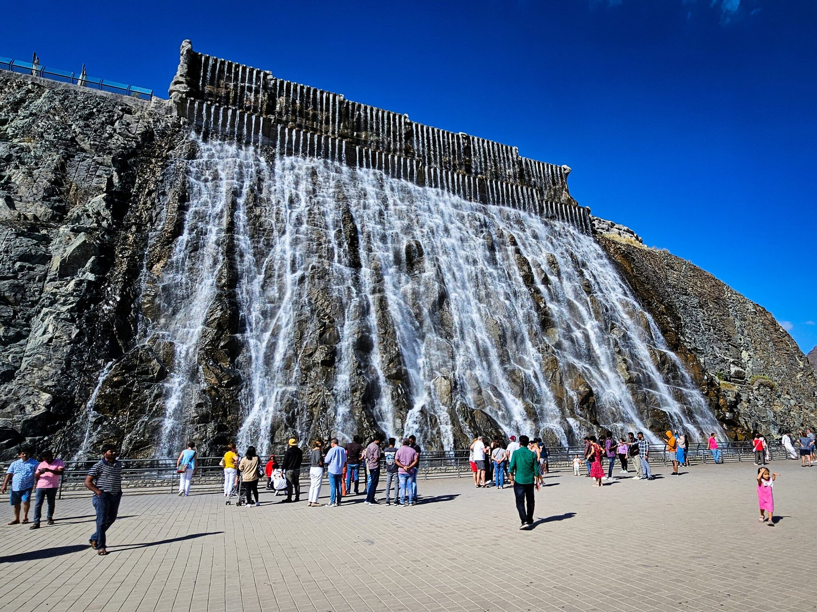 Khorfakkan's man-made waterfall cascading down rocky cliff face with visitors gathered at viewing area below against bright blue sky