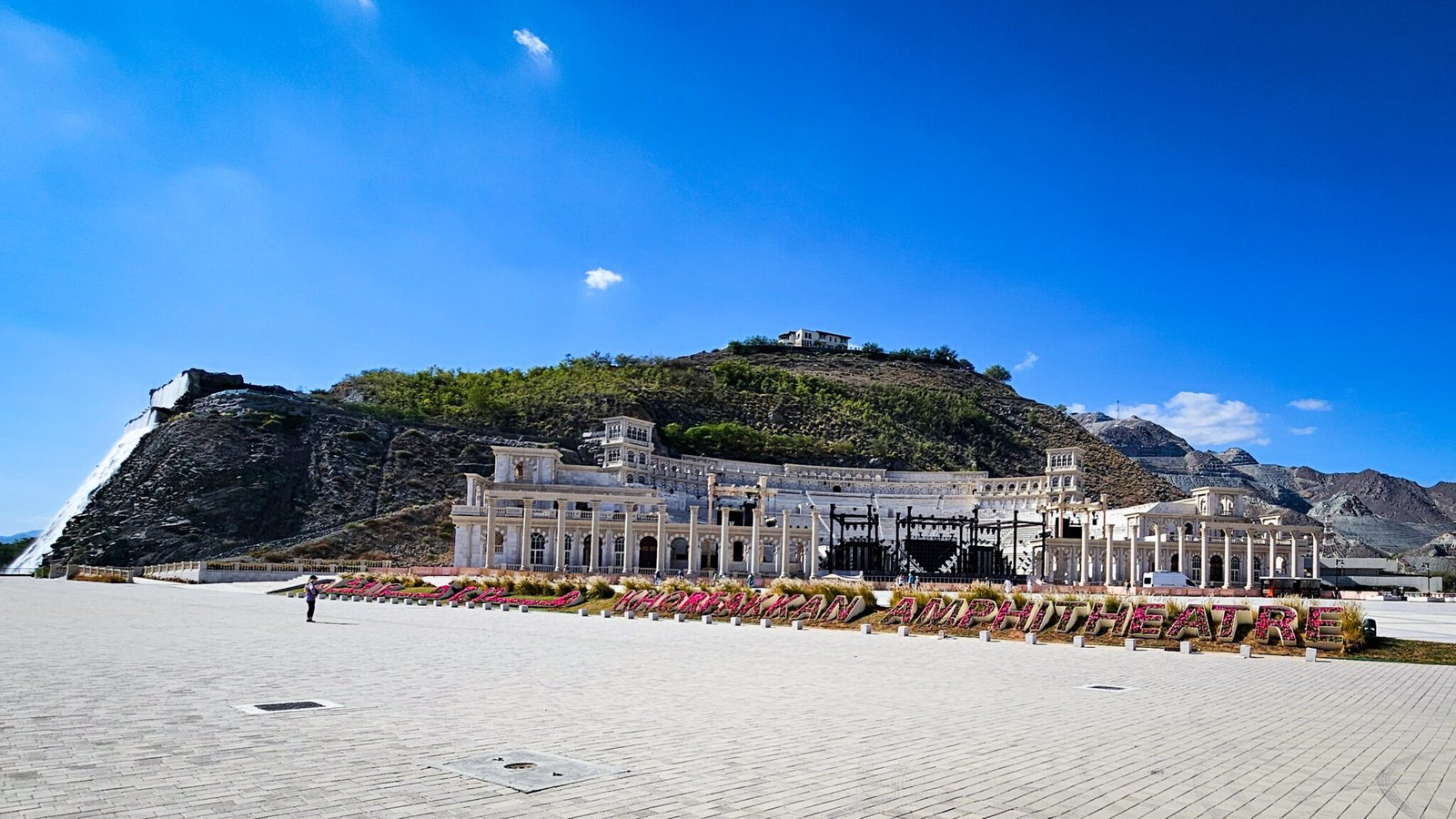 White Roman-inspired Khorfakkan Amphitheatre with floral landscaping nestled against rocky mountain backdrop under blue sky.