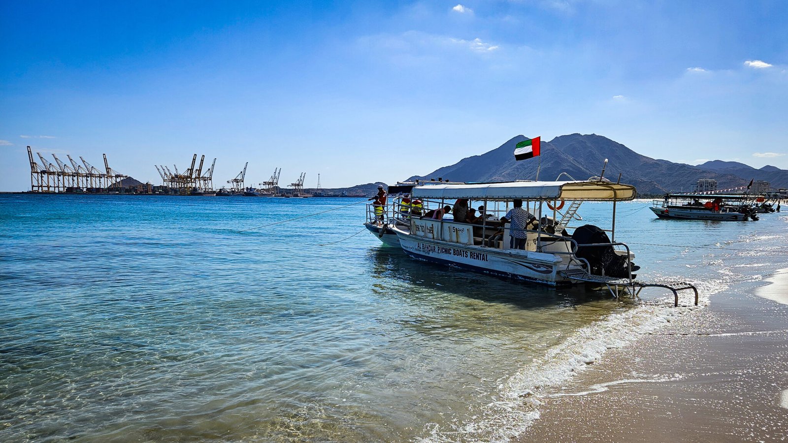 Tour boat with UAE flag on Khorfakkan beach shoreline with port cranes and Al Rabi mountain visible across the clear blue water of Khorfakkan Bay.
