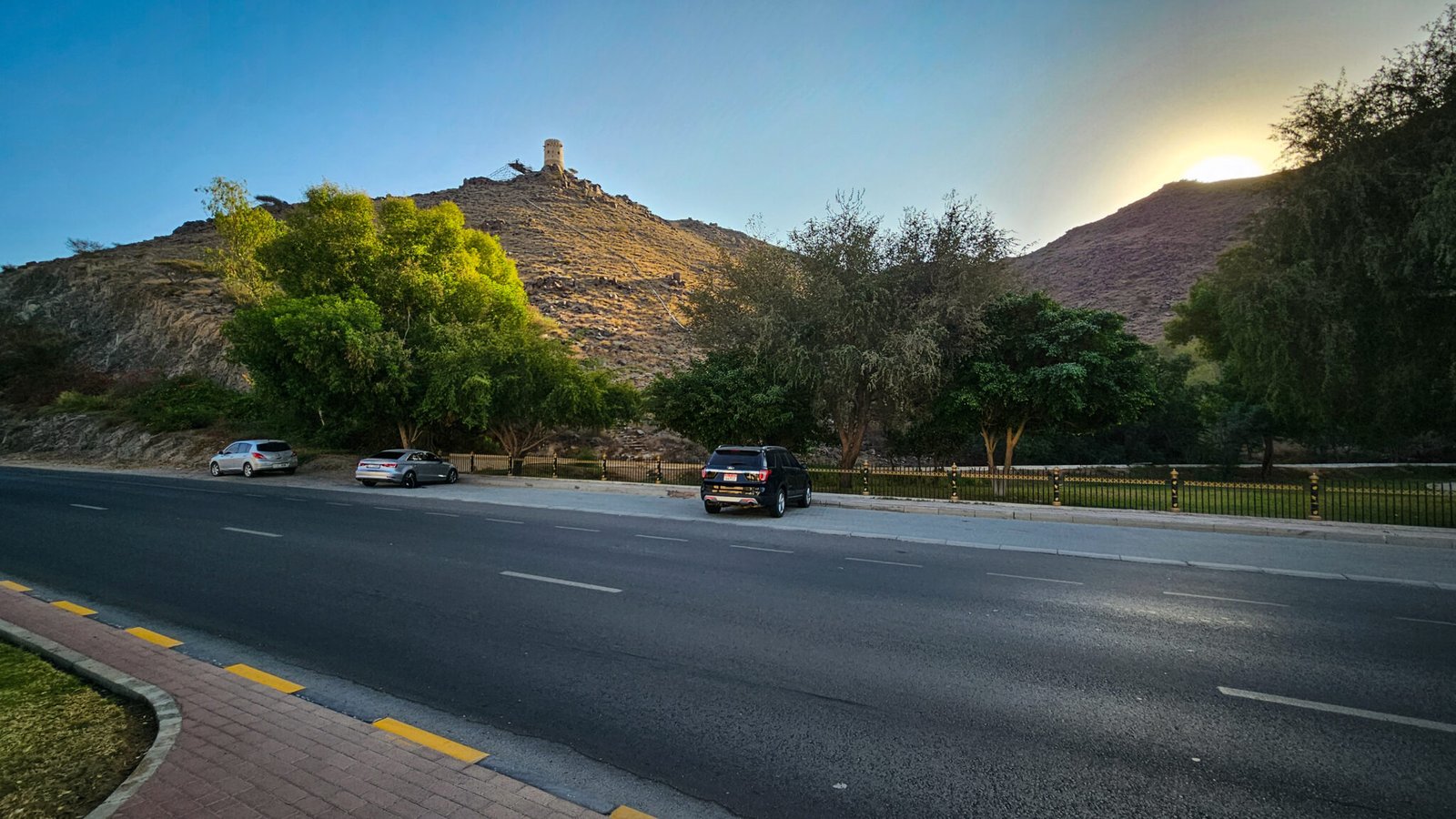 View of Al Suwaifa Park parking area in Khorfakkan with cars parked alongside road, green trees, and watchtower visible on hill marking the trailhead to Secret Beach.
