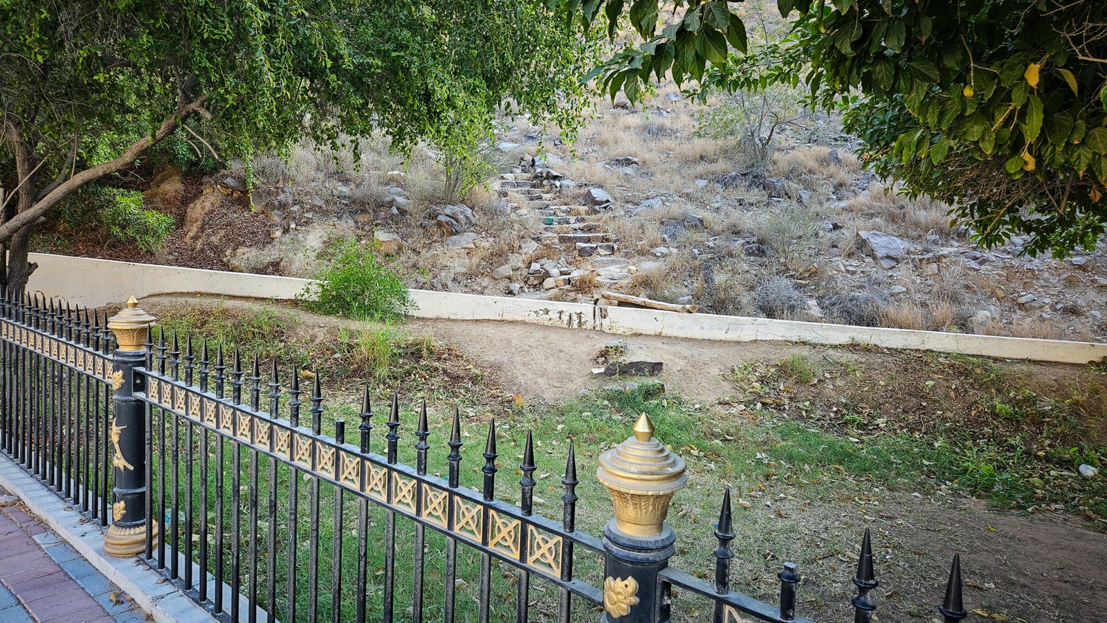 Stone steps marking the beginning of Secret Beach trail beyond the decorative black fence at Al Suwaifa Park, with shade trees overhead.