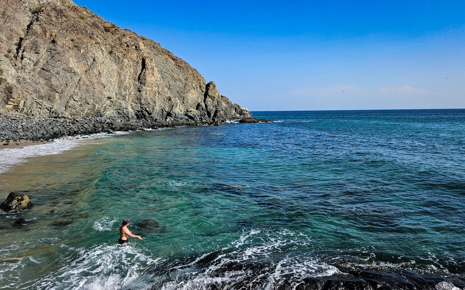 Person swimming in crystal-clear turquoise waters at secluded beach surrounded by dramatic rocky cliffs under blue sky.