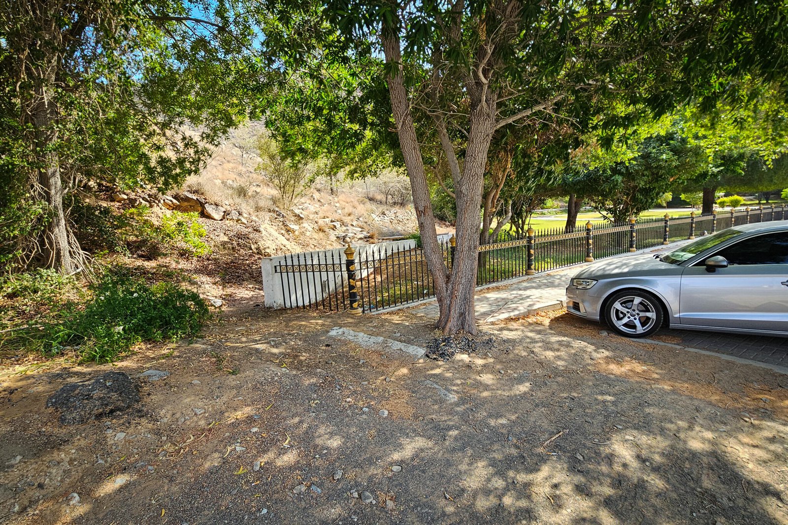 Hidden trailhead for Secret Beach path at Al Suwaifa Park showing dirt path around black fence with a silver car parked nearby and shade trees.