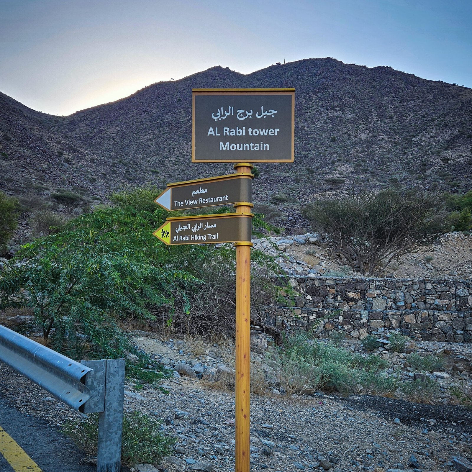 Wooden directional sign post showing Al Rabi Tower Mountain, Al Rabi Hiking Trail, and The View Restaurant with mountain backdrop in Khorfakkan.