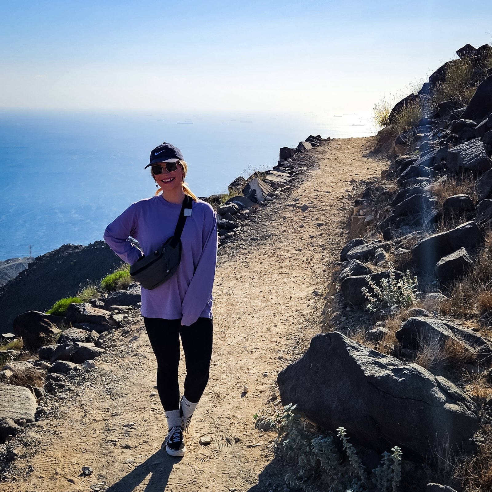 Female hiker in purple top and cap walking along the Al Rabi mountain trail with panoramic Gulf of Oman views and distant cargo ships visible