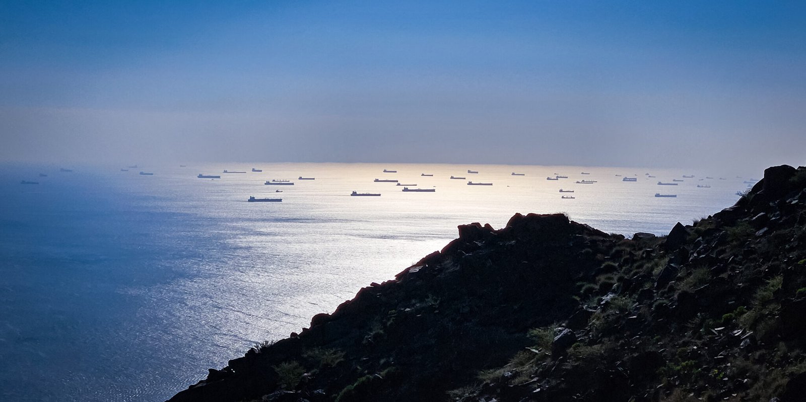 Dozens of cargo ships anchored in formation on the Gulf of Oman as seen from Al Rabi Mountain with sunlight reflecting off the water.