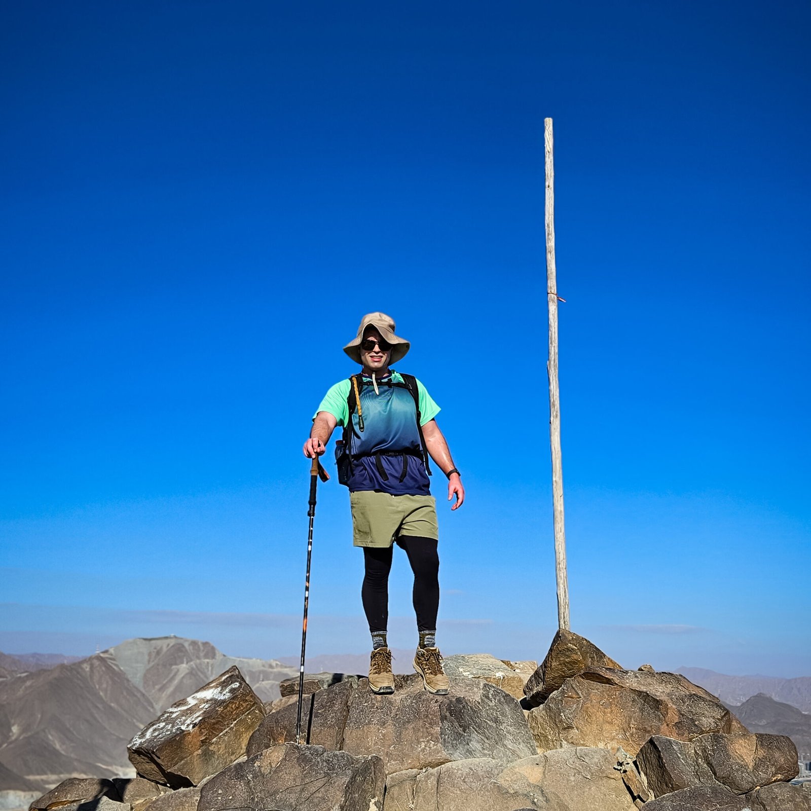 Hiker standing on rocky Al Shahs peak on Al Rabi mountain with trekking pole, wide-brimmed hat, and technical hiking clothing against blue sky and Hajar Mountains backdrop.