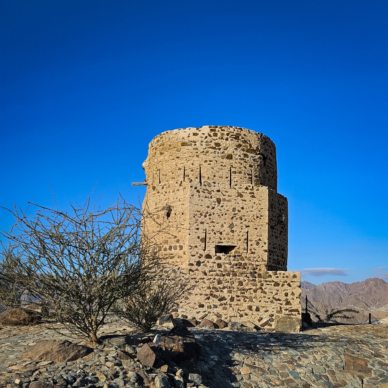 Ancient stone Al Rabi Watchtower against deep blue sky with desert shrub and Hajar Mountains visible in the background.