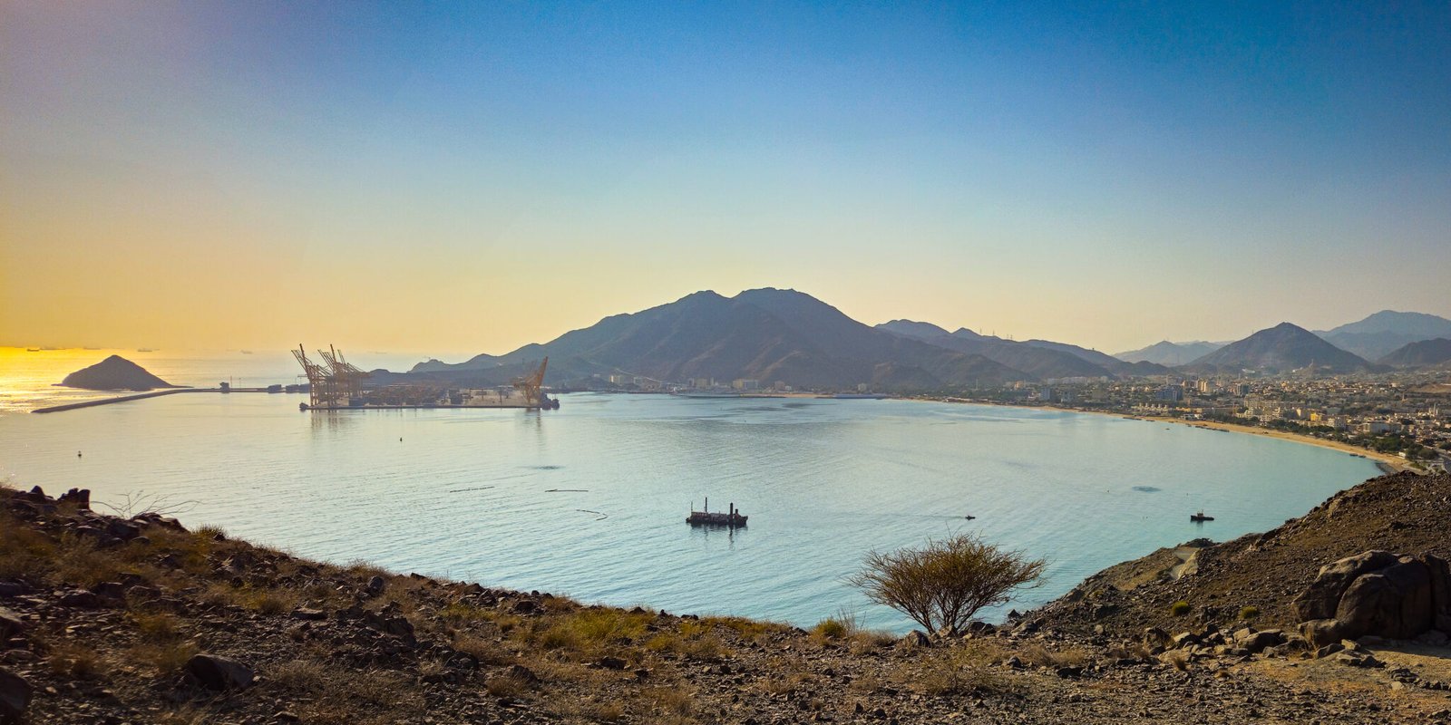 Panoramic view of Khorfakkan Bay with Al Rabi mountain, Khorfakkan Port facilities, and Shark Island visible from elevated hiking trail viewpoint.