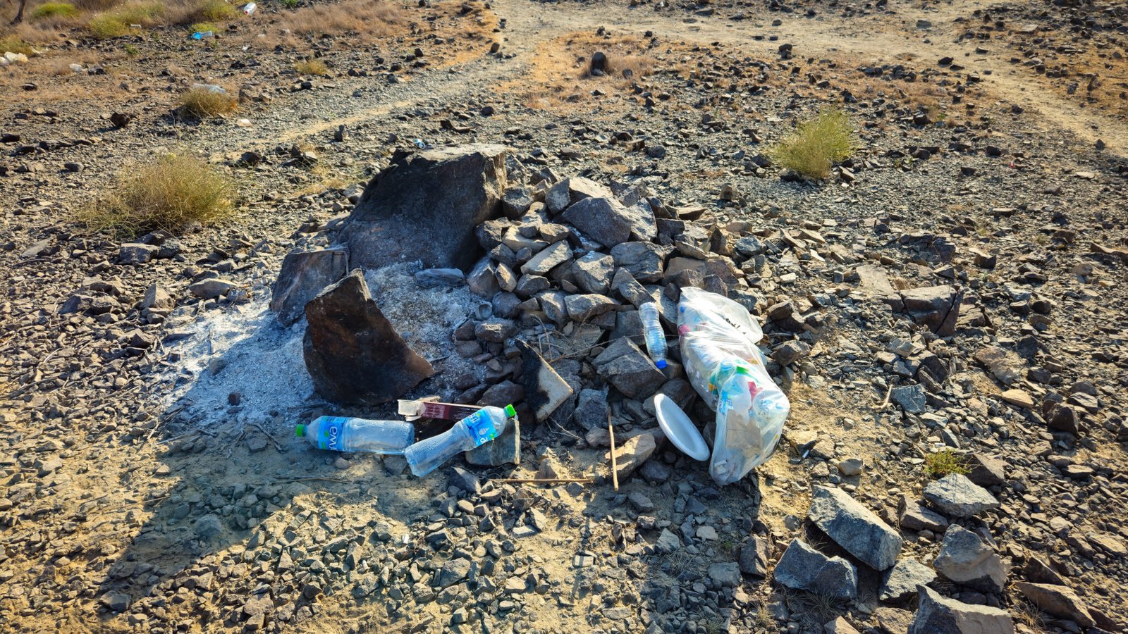 Pile of trash including plastic bottles, food containers, and garbage bags left scattered around makeshift fire pit on mountain trail.