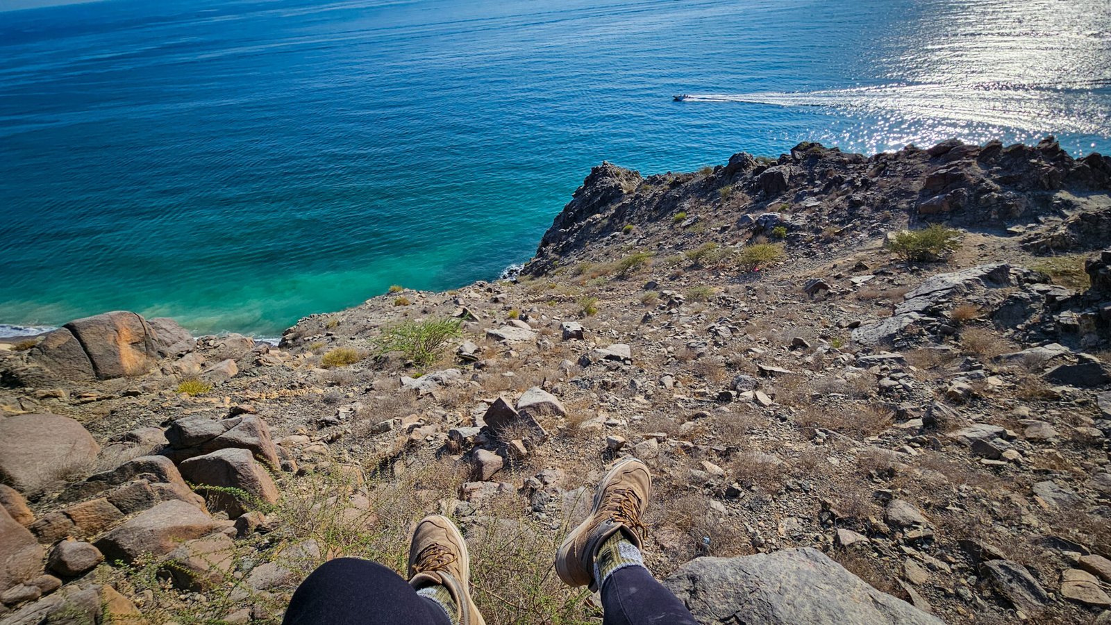 First-person view of hiker's boots resting on rocky cliff edge overlooking turquoise Gulf of Oman waters with a boat visible in the distance.