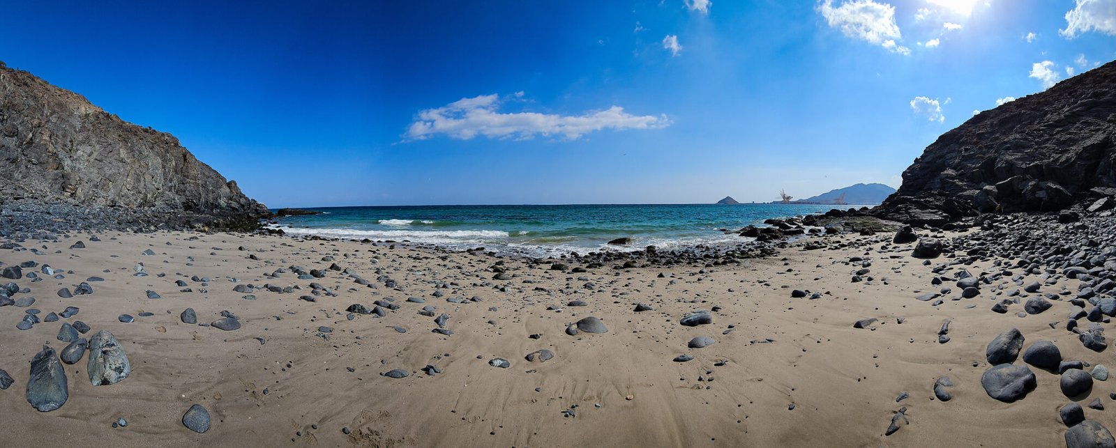 Pristine sandy beach with scattered rocks enclosed by dramatic cliffs, showing Al Rabi Mountain, port facilities, and Shark Island across the bay.