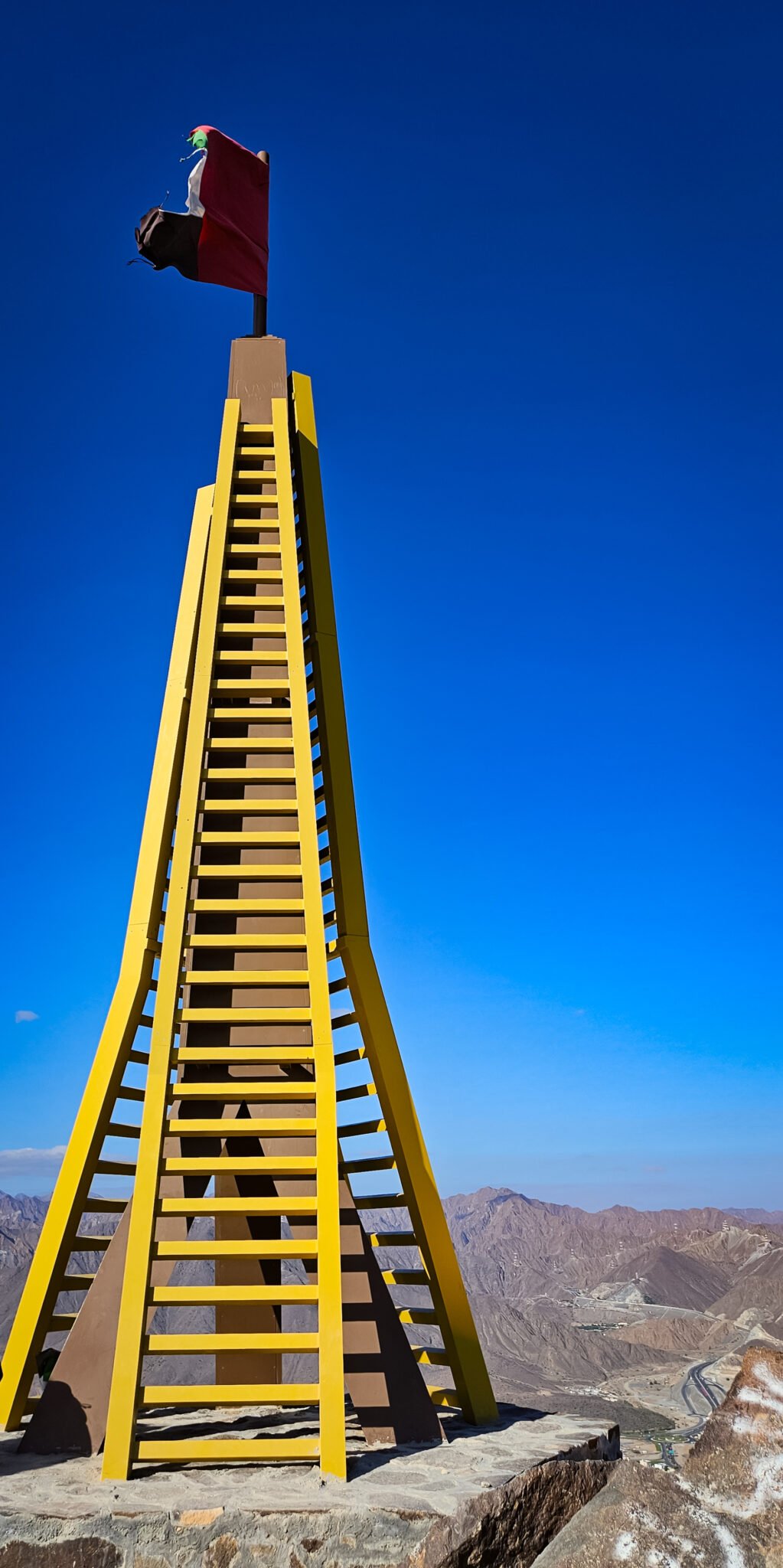 Tall yellow ladder-like structure at Al Rabi Mountain summit with tattered UAE flag against brilliant blue sky and Hajar Mountains panorama.